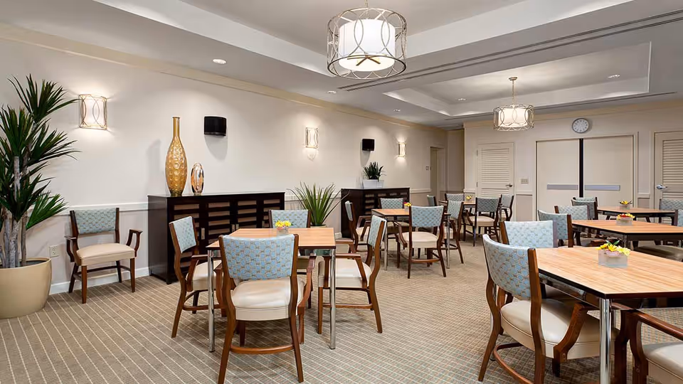 A well-lit dining room with multiple wooden tables and cushioned chairs arranged neatly. The room features beige walls, carpeted flooring with a striped pattern, decorative plants, and modern light fixtures hanging from the ceiling. There are cabinets along the wall with decorative vases and small plants on top.