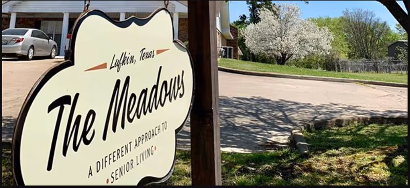 Sign reading The Meadows at the entrance to a senior living facility with a driveway, parked car, and trees