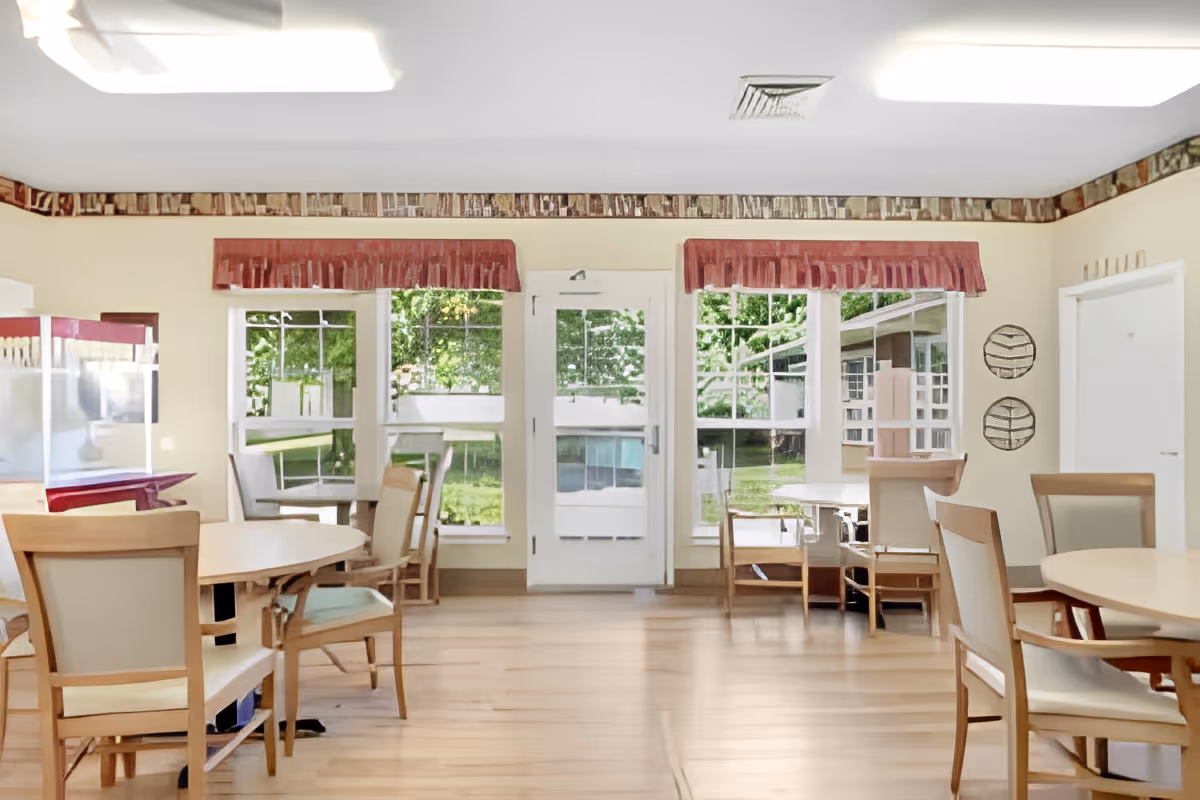A bright dining room with multiple round tables and wooden chairs with light-colored cushions. Large windows and a glass door provide a view of green outdoor trees and grass. The room has light-colored walls with a decorative border near the ceiling and red valances above the windows. The floor is wooden, and there is a popcorn machine on the left side.