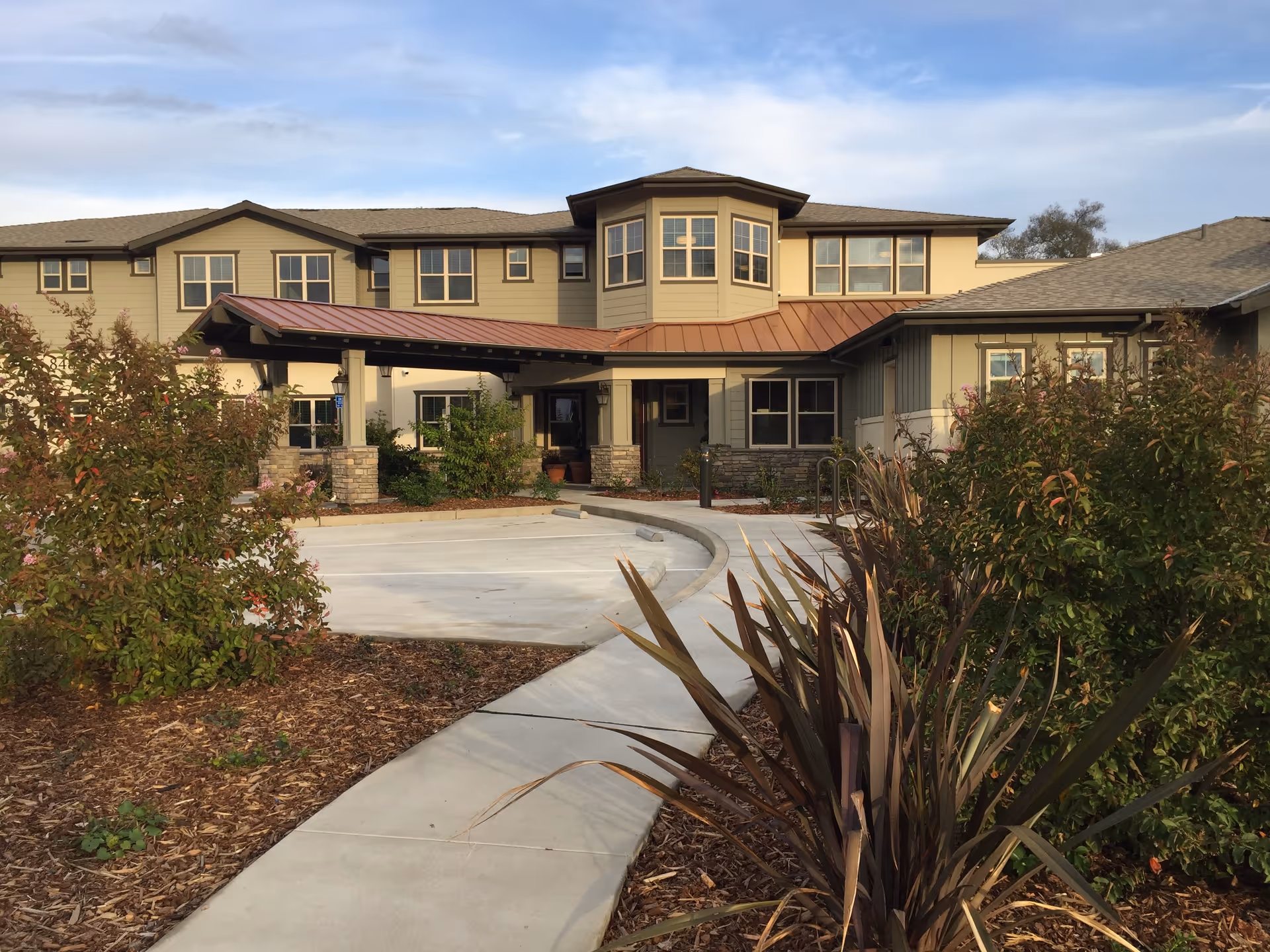 Exterior view of a two-story senior living facility building with beige siding and a brown roof. There is a covered entrance supported by stone pillars, a curved concrete walkway leading to the entrance, and landscaped bushes and plants around the pathway.