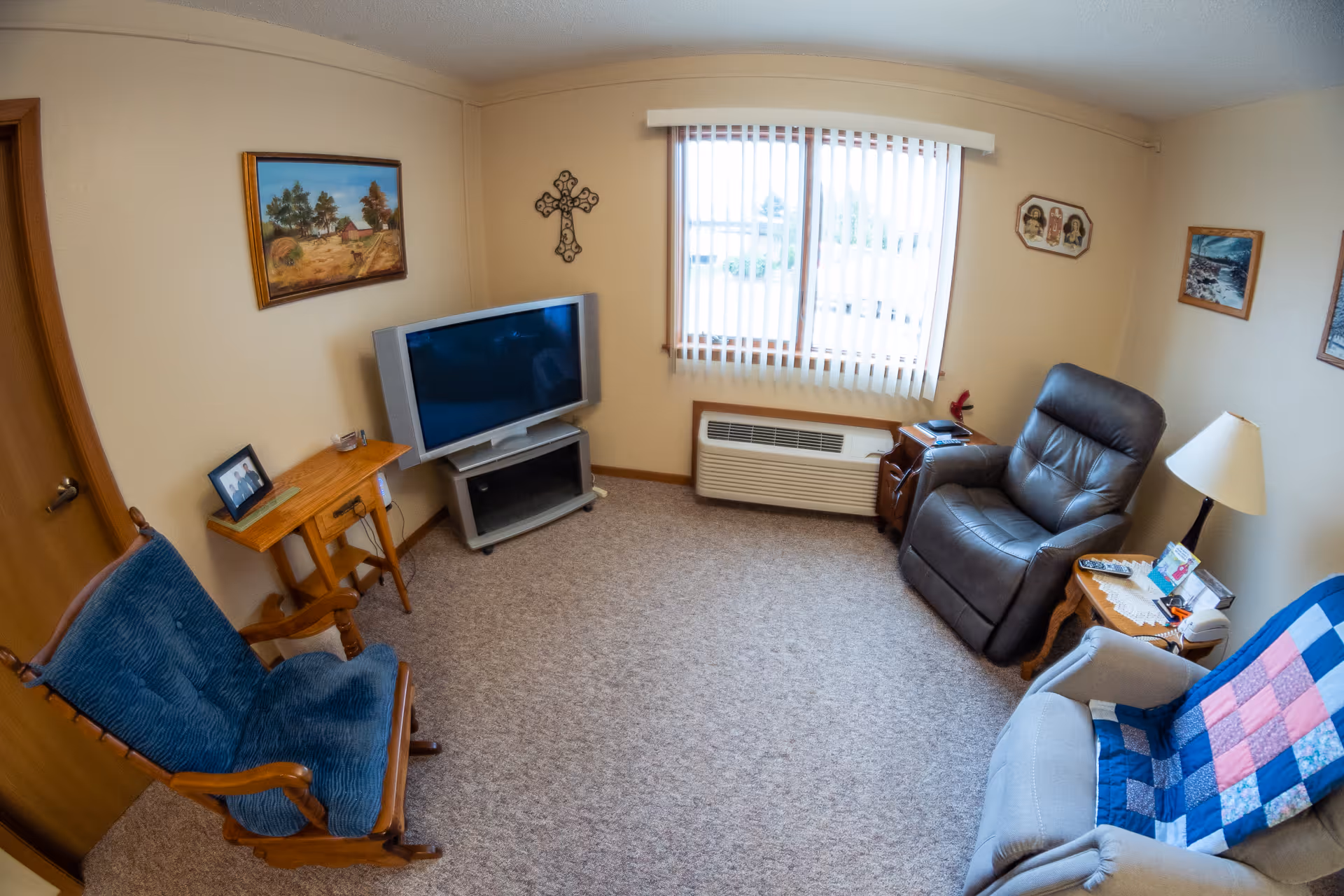 A cozy living room in an assisted living facility featuring a large window with vertical blinds, a flat-screen TV on a stand, a wooden rocking chair with a blue cushion, a dark leather recliner, a light-colored armchair with a blue and pink patchwork quilt draped over it, a wooden side table with a lamp and various items, and framed artwork and a decorative cross on the beige walls.