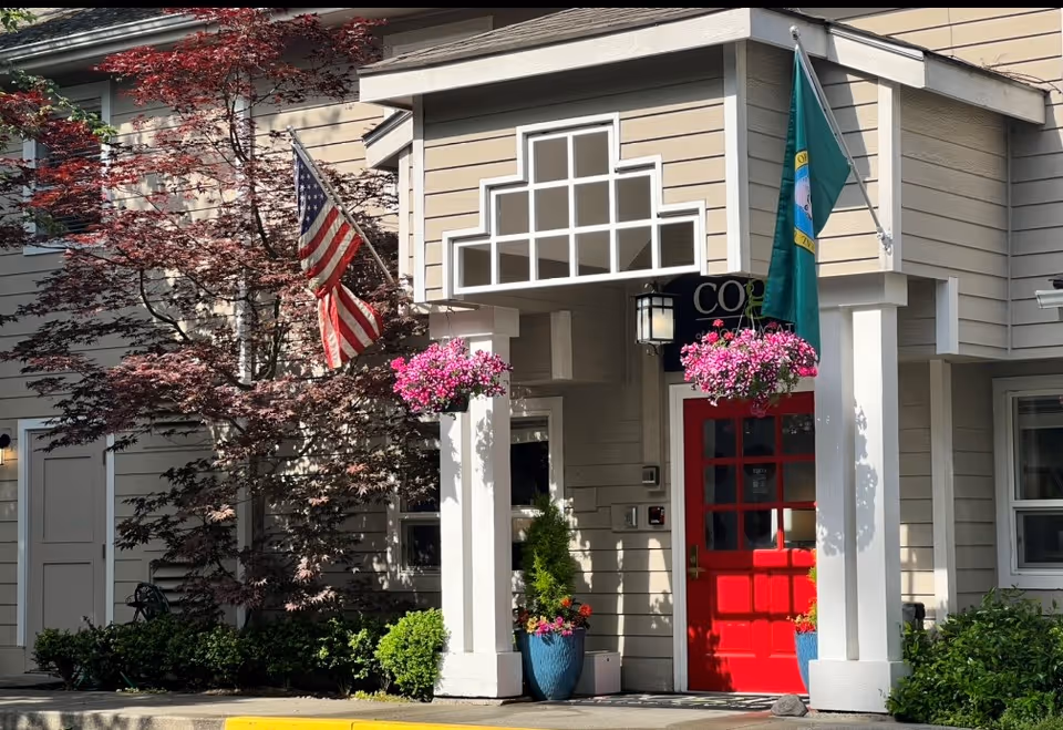 Beige building entrance with a bright red door, two flags, hanging pink flower baskets, and potted plants.