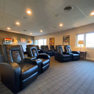A common area with multiple black leather recliner chairs arranged in rows facing the same direction. The room has carpeted flooring, beige walls, and a ceiling with recessed lighting. There are two windows with table lamps on side tables beneath them, and framed artwork on the walls. A popcorn machine is visible in the back corner of the room.