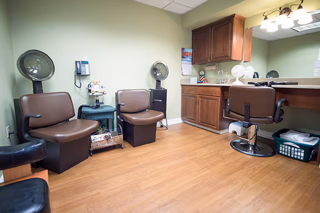 Interior of a hair salon area in a senior living facility with three brown salon chairs, two hair dryers mounted on stands, a small table with a teddy bear, a wall-mounted phone, wooden cabinets with a countertop and sink, and a laundry basket on the floor.
