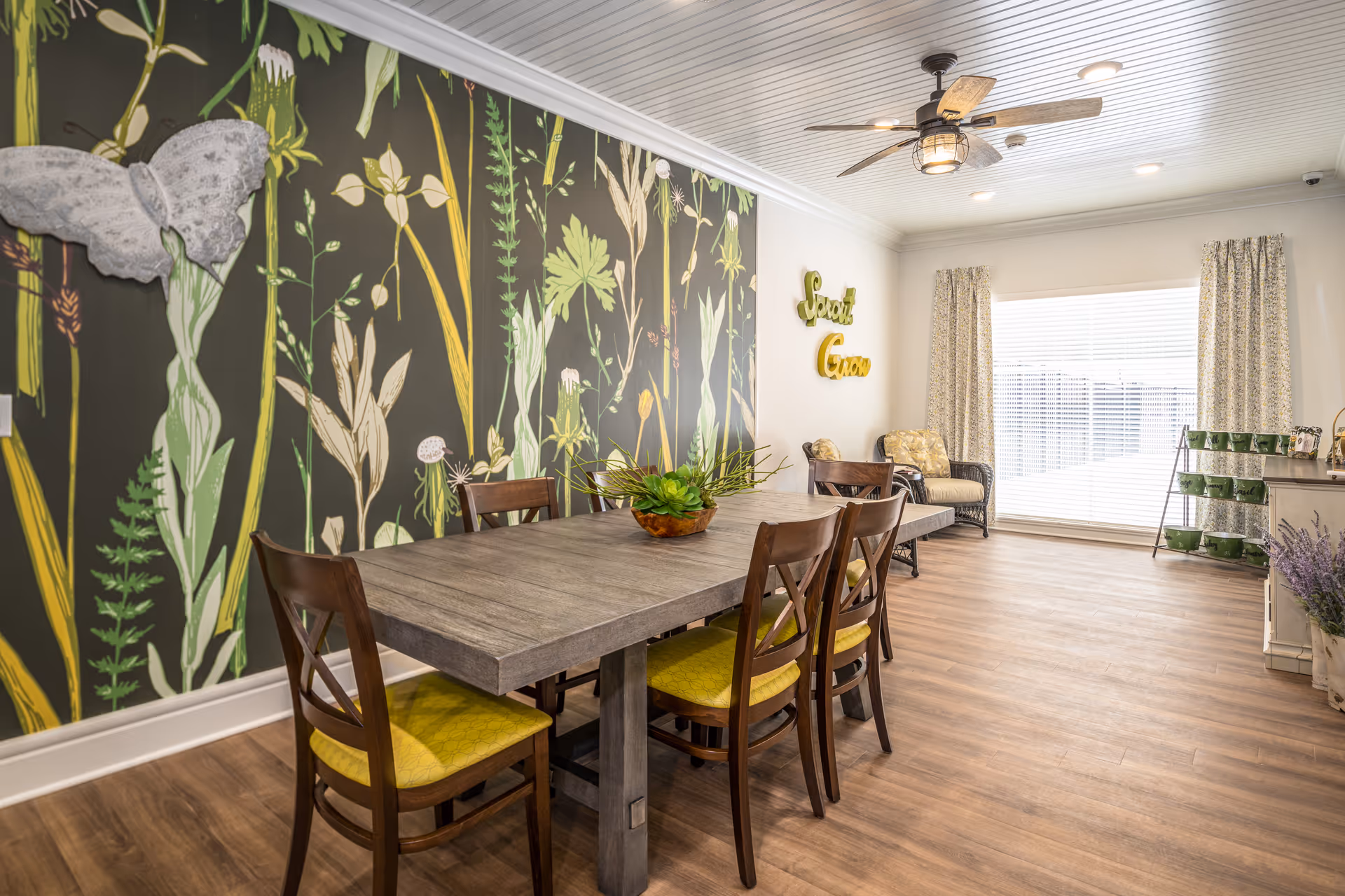Communal dining room featuring a long wooden table with six chairs, a botanical accent wall, ceiling fan, and a large window letting in natural light.