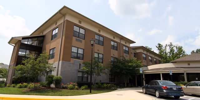 Exterior view of a three-story senior living facility building with brick and beige siding, surrounded by landscaped greenery and a parking area with cars parked near the entrance under a partly cloudy sky.