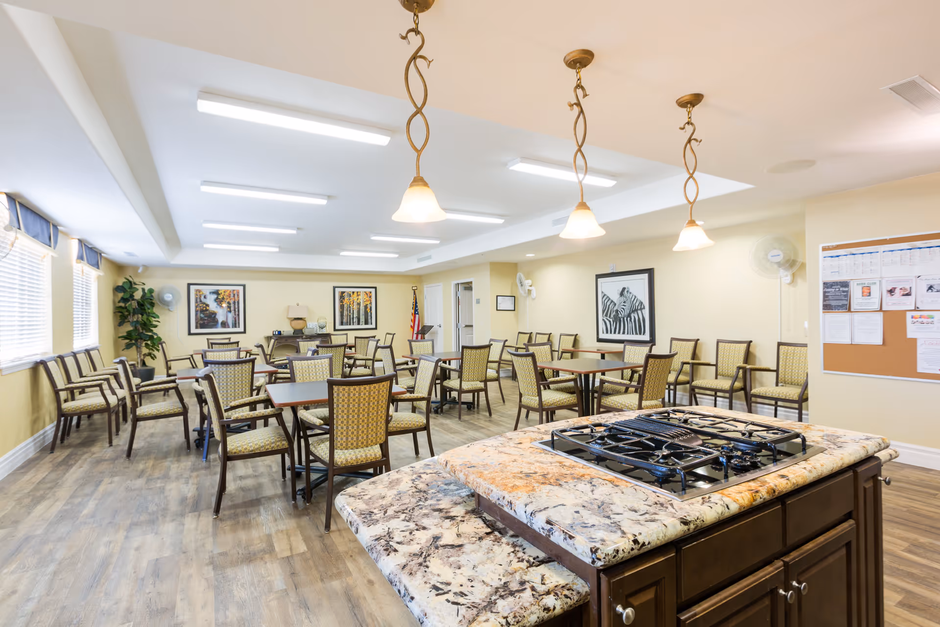 A bright and spacious dining area with multiple tables and chairs arranged neatly. The room features large windows with blinds, framed artwork on the walls, and a kitchen island with a stove in the foreground. Pendant lights hang from the ceiling, and a bulletin board is visible on one wall.