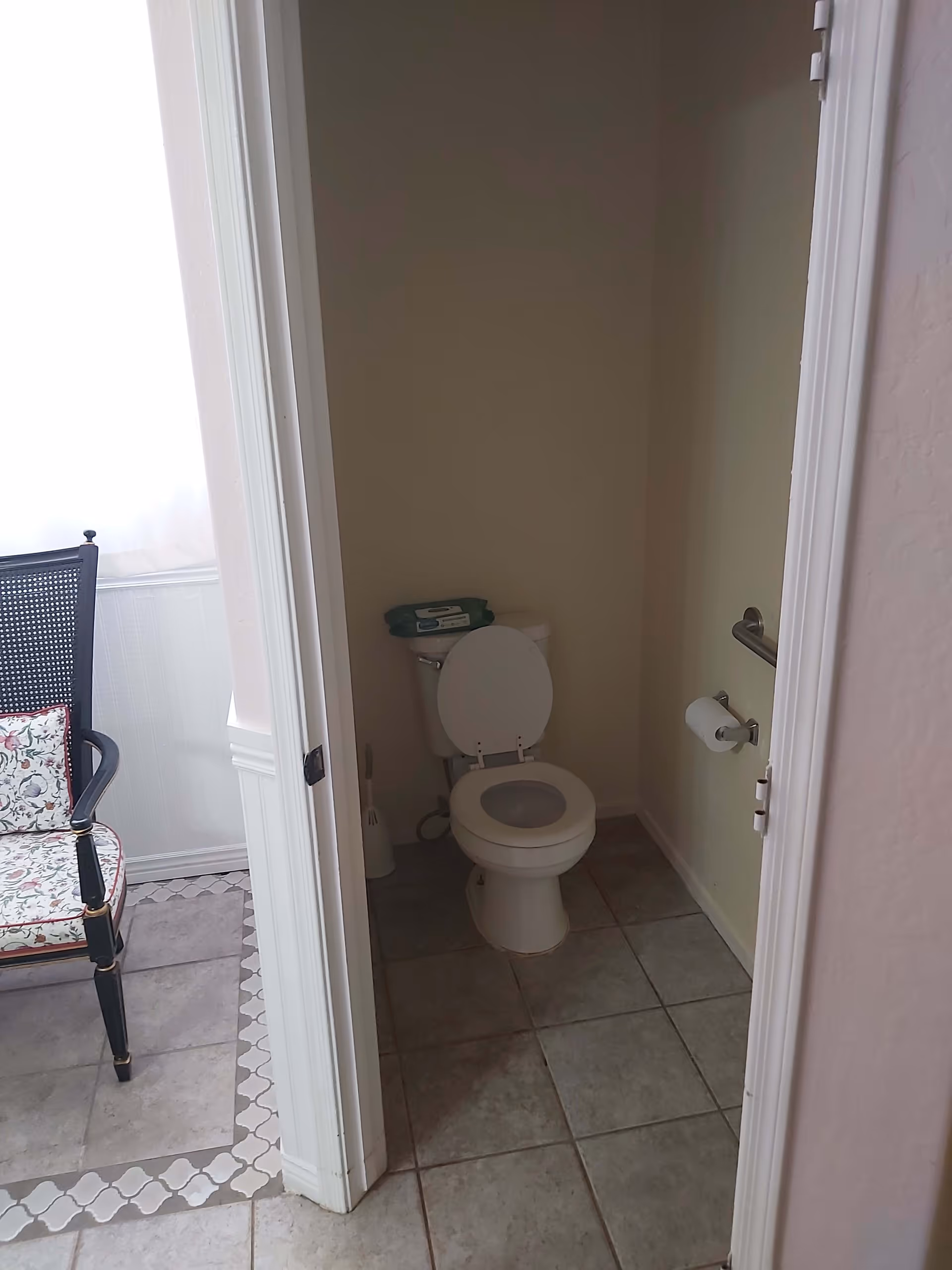 A small bathroom with a white toilet that has the seat and lid raised. There is a toilet paper holder mounted on the wall to the right and a toilet brush on the floor to the left. The bathroom floor is tiled in gray. Adjacent to the bathroom is a room with a black chair that has a floral cushion, and the floor has a decorative tile border.
