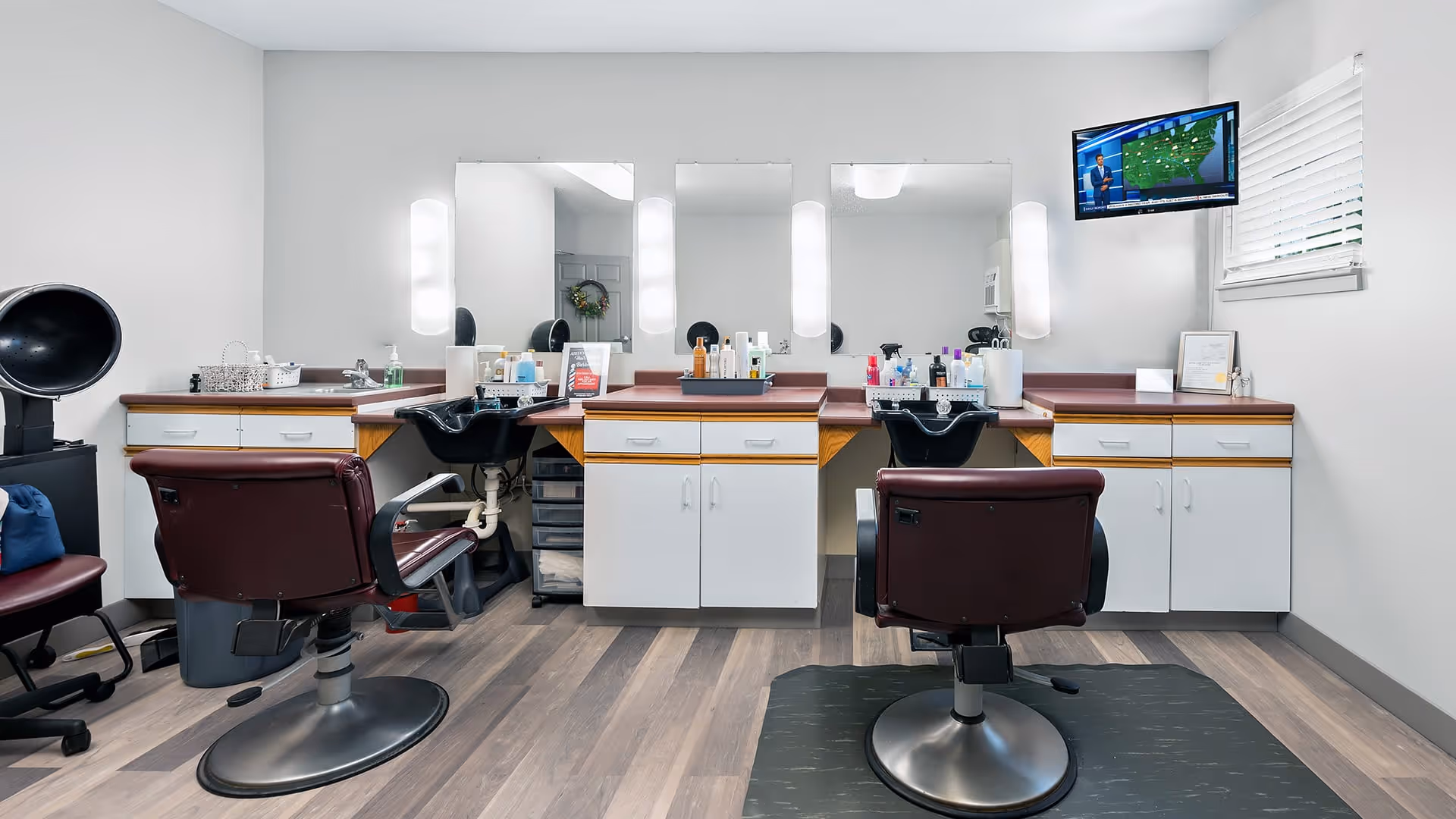 Interior view of a salon area with two maroon salon chairs facing mirrors and sinks. The room has white walls, wooden flooring, and white cabinets with brown countertops. A TV is mounted on the wall showing a weather forecast, and various hair care products are arranged on the countertops.