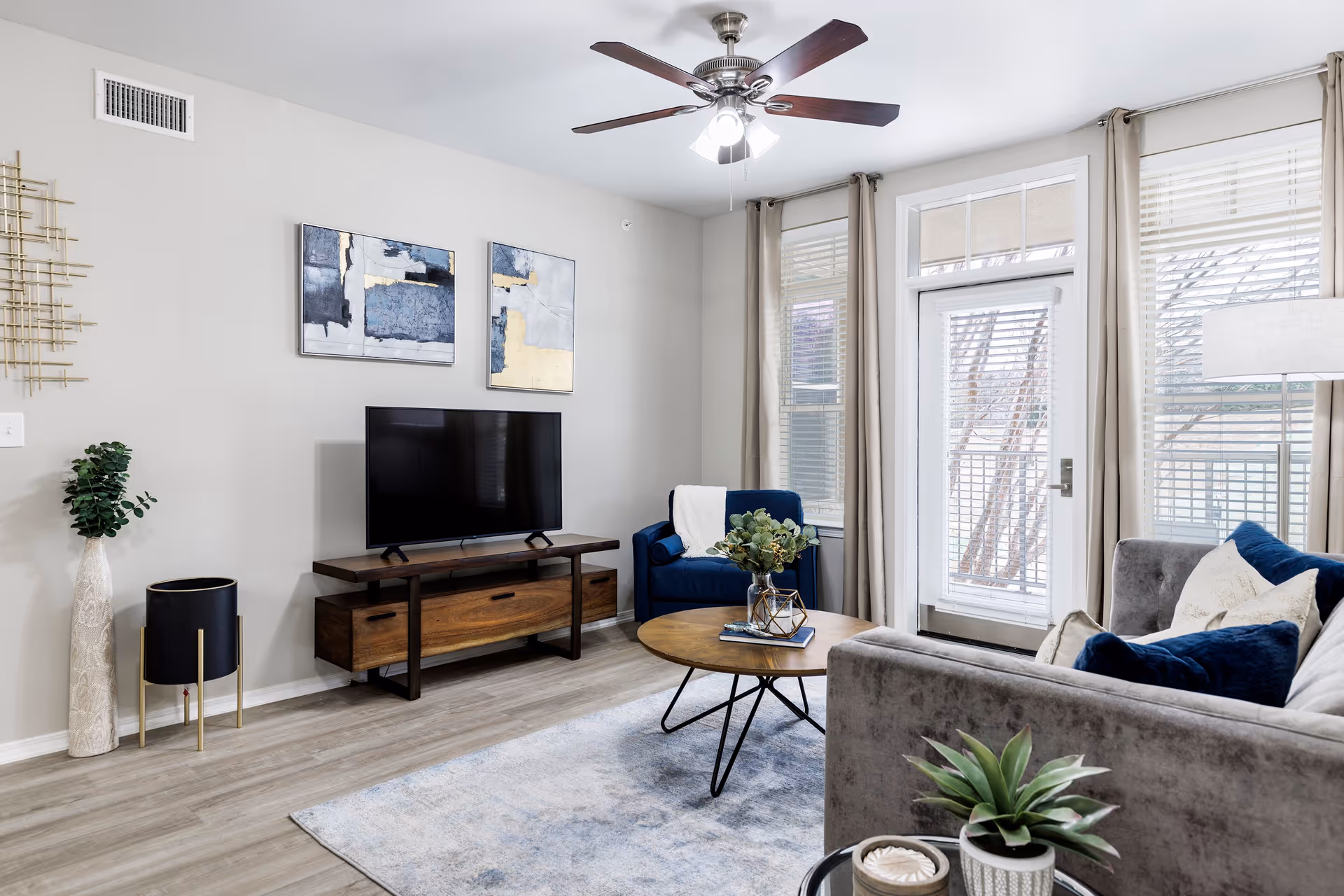 Well-lit modern living room with a sofa and armchair, TV on a wooden console, round coffee table and a glass door to a balcony.