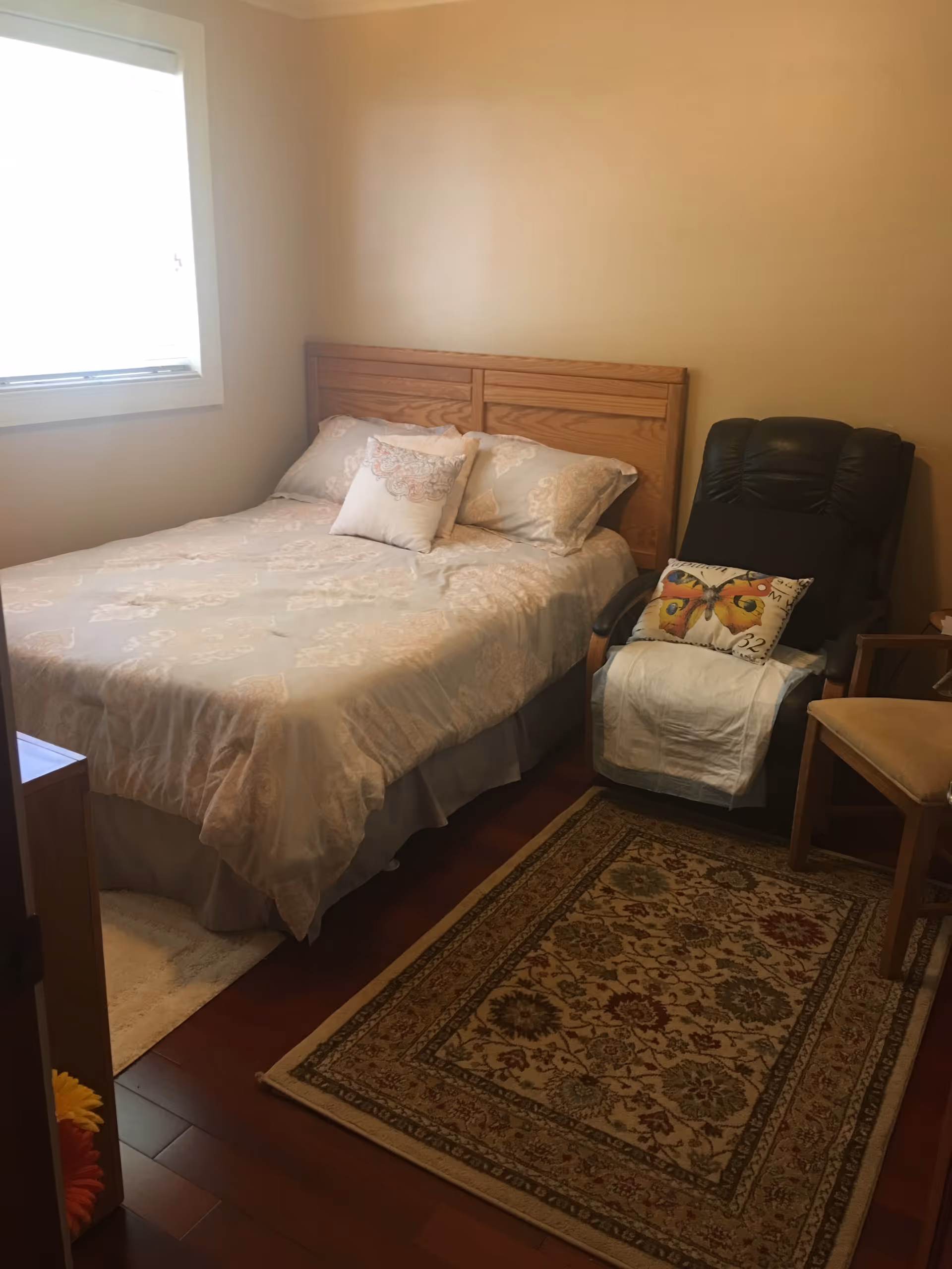 A cozy bedroom with a wooden headboard bed covered in light-colored patterned bedding and pillows. Next to the bed is a black recliner chair with a butterfly pillow and a white cloth draped over the seat. There is also a wooden chair and a patterned area rug on the wooden floor. A window with a white blind is on the left wall.