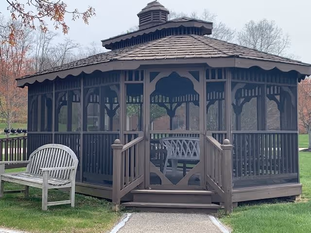 A brown wooden gazebo with a shingled roof and screened sides, situated on a grassy area with a wooden bench nearby. Trees with sparse leaves are visible in the background under an overcast sky.