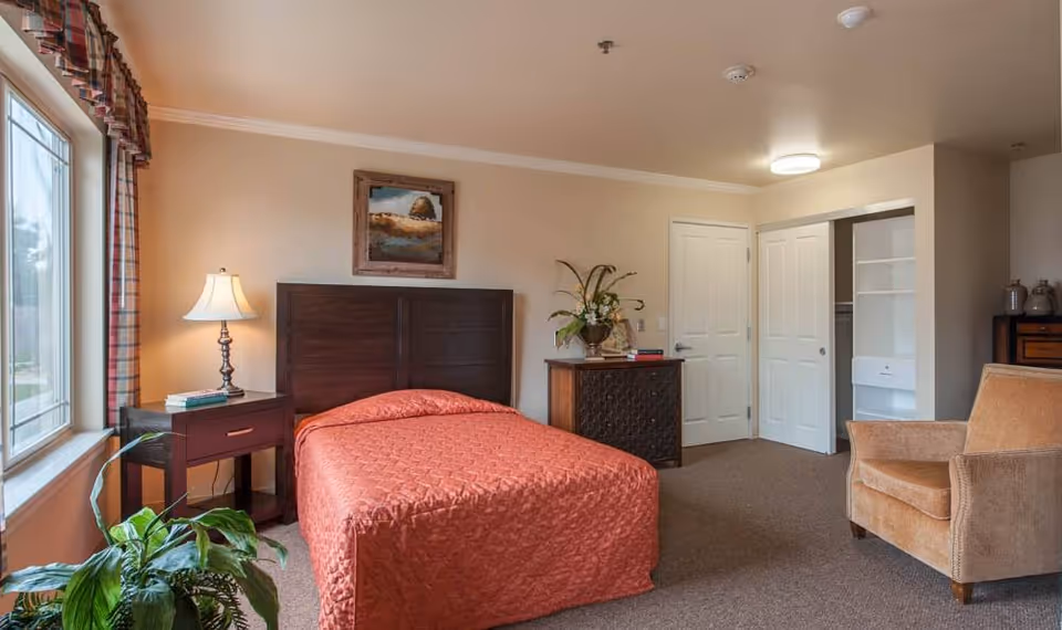 A senior living bedroom with a large window on the left, a bed with an orange quilted bedspread, a wooden headboard, a nightstand with a lamp and books, a plant near the window, a dresser with a flower arrangement, an armchair, and an open closet with white shelving.