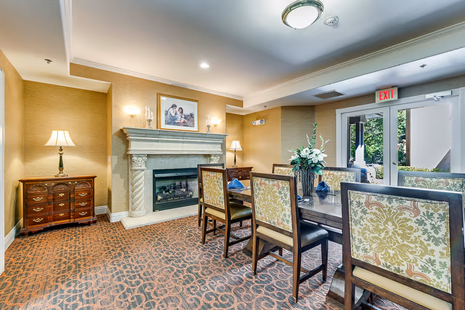 A cozy dining area in a senior living facility with a wooden dining table surrounded by six upholstered chairs featuring a floral pattern. The room has a patterned carpet, a decorative fireplace with a framed picture above it, two table lamps on wooden side tables, and a vase with flowers on the table. There is a glass door leading outside with an exit sign above it.