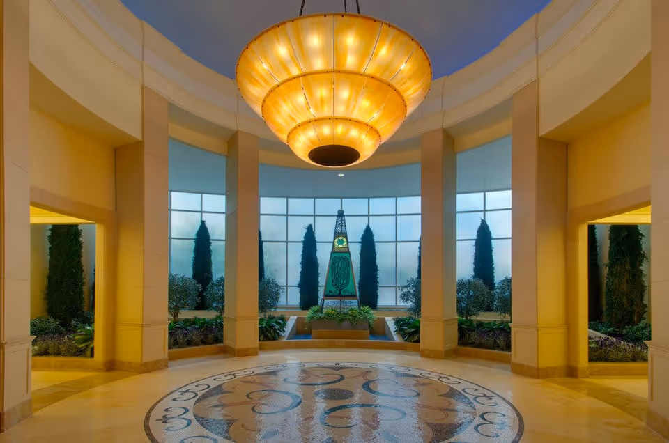 A spacious, elegant interior area with a large circular mosaic floor design, tall columns, and a grand, warm-toned chandelier hanging from the ceiling. The background features tall windows with frosted glass, flanked by tall green plants and an artistic obelisk centerpiece surrounded by greenery.