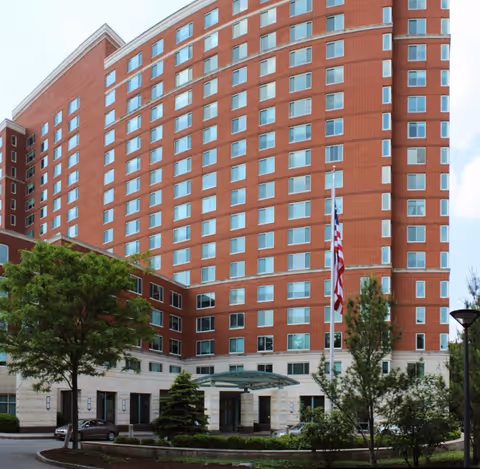 Exterior view of a large multi-story red brick building with many windows, an entrance canopy, trees, and an American flag on a flagpole in front of the building.
