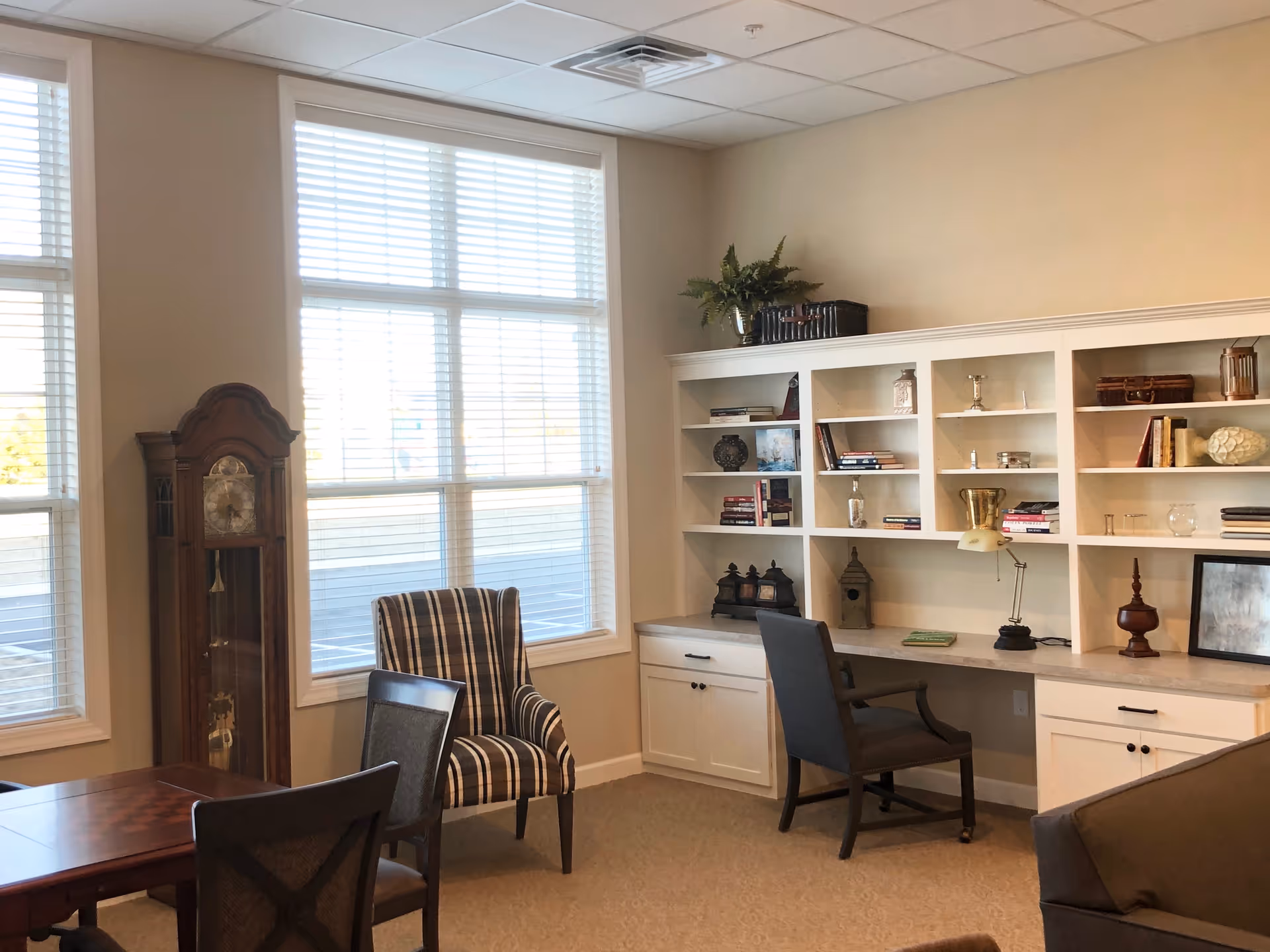 A cozy senior living common area with large windows letting in natural light. The room features a tall grandfather clock, a striped armchair, a wooden table with chairs, and a built-in white shelving unit with various decorative items and books. A desk with a chair and a lamp is integrated into the shelving unit.