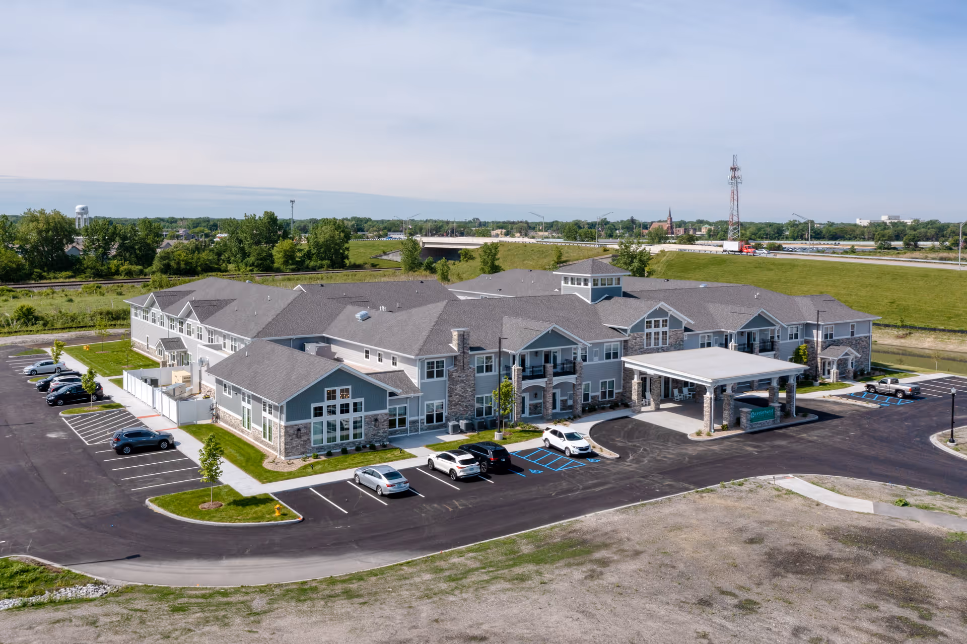 Aerial view of Cedarhurst Senior Living of Dyer, a large two-story senior living facility with gray roofs and stone accents. The building is surrounded by a parking lot with several cars parked, green lawns, and trees. In the background, there is a highway, a water tower, and a communication tower under a partly cloudy sky.