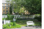 Outdoor garden area with green grass, trees, and white wooden benches. A white picket fence separates the garden from a yellow multi-story building in the background.