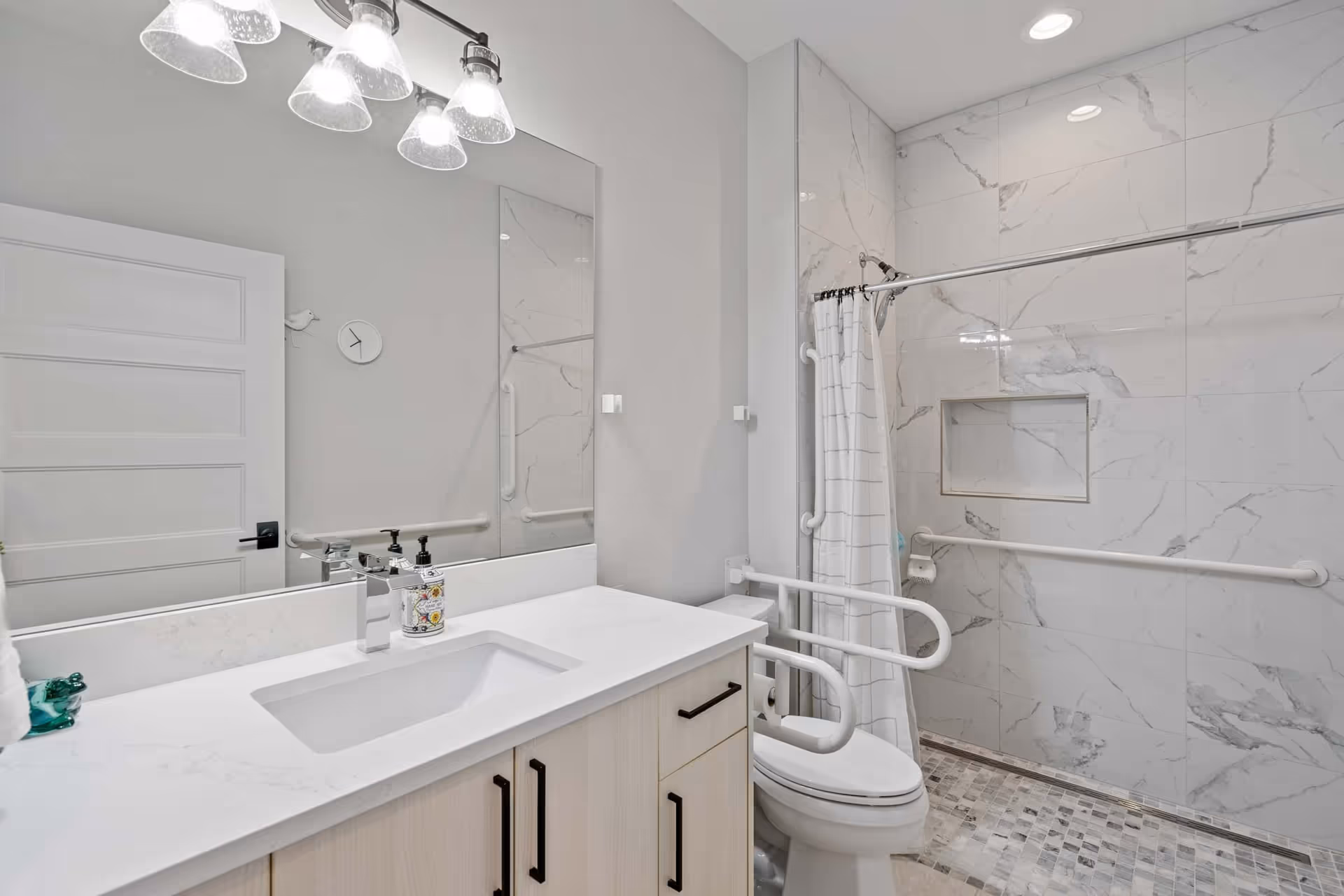 A modern bathroom with a white countertop sink, light wood cabinetry, and a large mirror above. The bathroom features a toilet with safety grab bars on both sides and a walk-in shower with marble-patterned tiles, a built-in niche, and a shower curtain. The walls are light-colored, and there are multiple light fixtures above the mirror providing bright illumination.