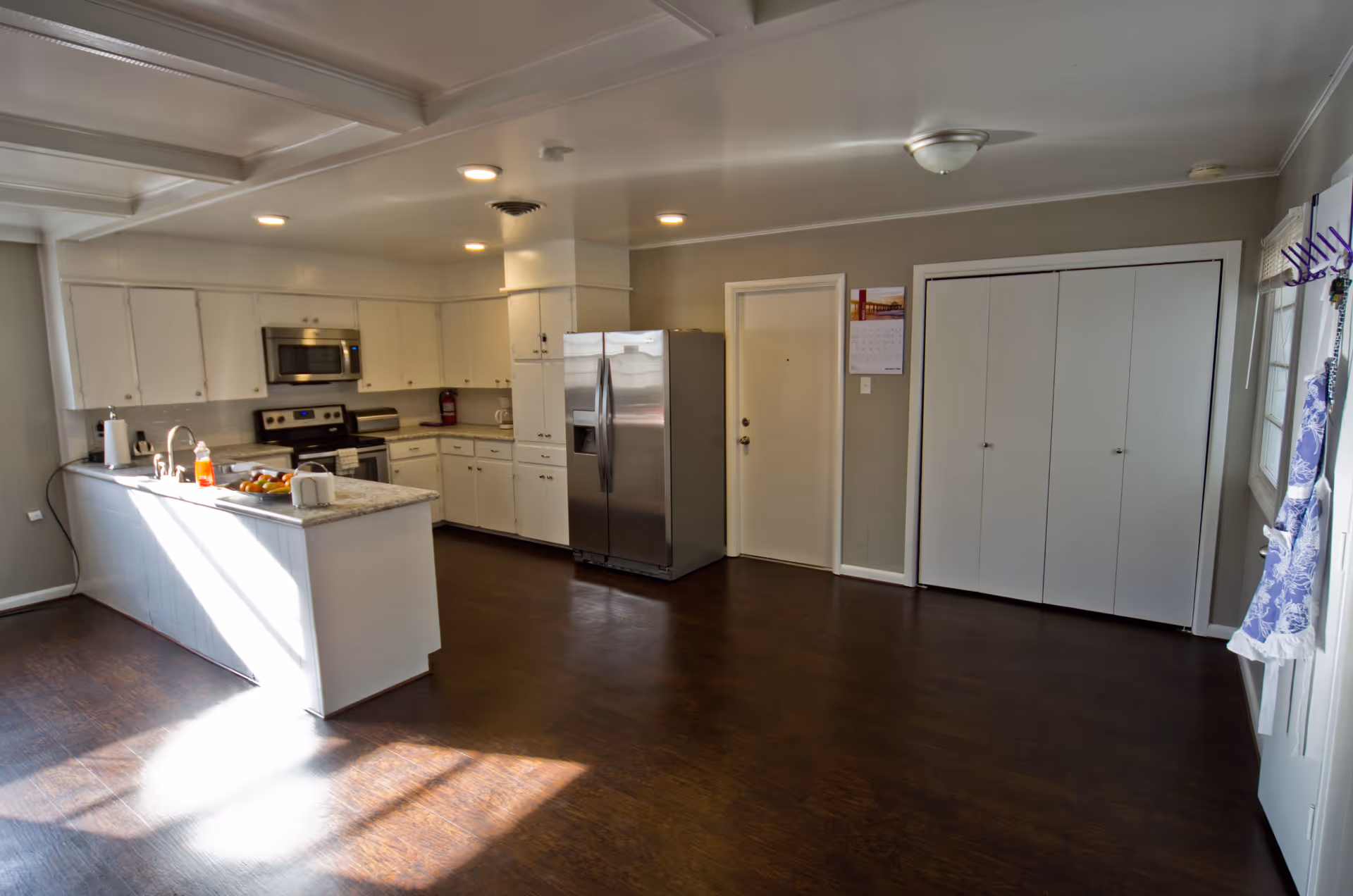 A spacious kitchen with white cabinets, a stainless steel refrigerator, stove, and microwave. There is a kitchen island with a sink and some items on the counter including a bowl of fruit and a soap dispenser. The floor is dark wood, and there is a door and a closet with white doors on the far wall. A calendar is hanging on the wall next to the door.