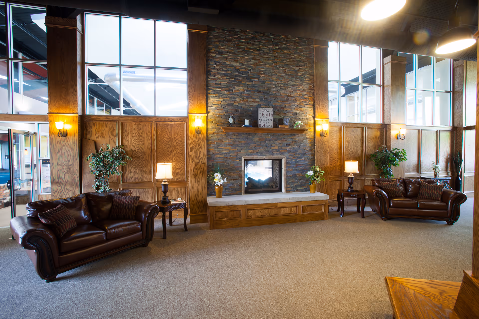 A spacious and well-lit living room area with two dark brown leather sofas facing each other, a stone fireplace in the center with a wooden mantel, and large windows letting in natural light. The room features wooden paneling on the walls, several lamps on side tables, and decorative plants.
