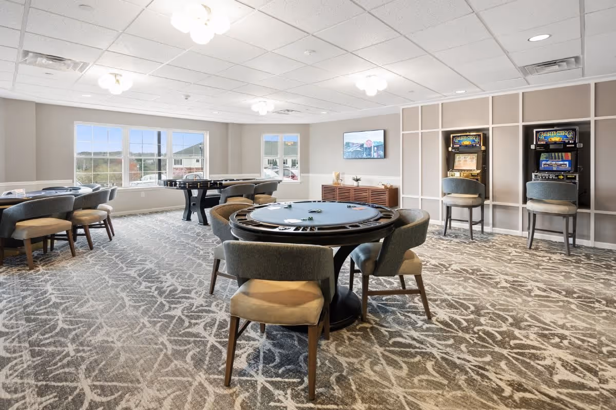A bright and spacious game room with large windows letting in natural light. The room features multiple round tables with chairs, a foosball table, two slot machines against the wall, and a flat-screen TV mounted on the wall. The carpet has a patterned design, and the walls are painted in neutral tones.