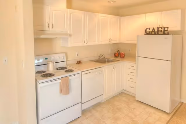 A compact white kitchen with an electric stove, dishwasher, sink, refrigerator and upper cabinets with 'CAFE' letters on top.