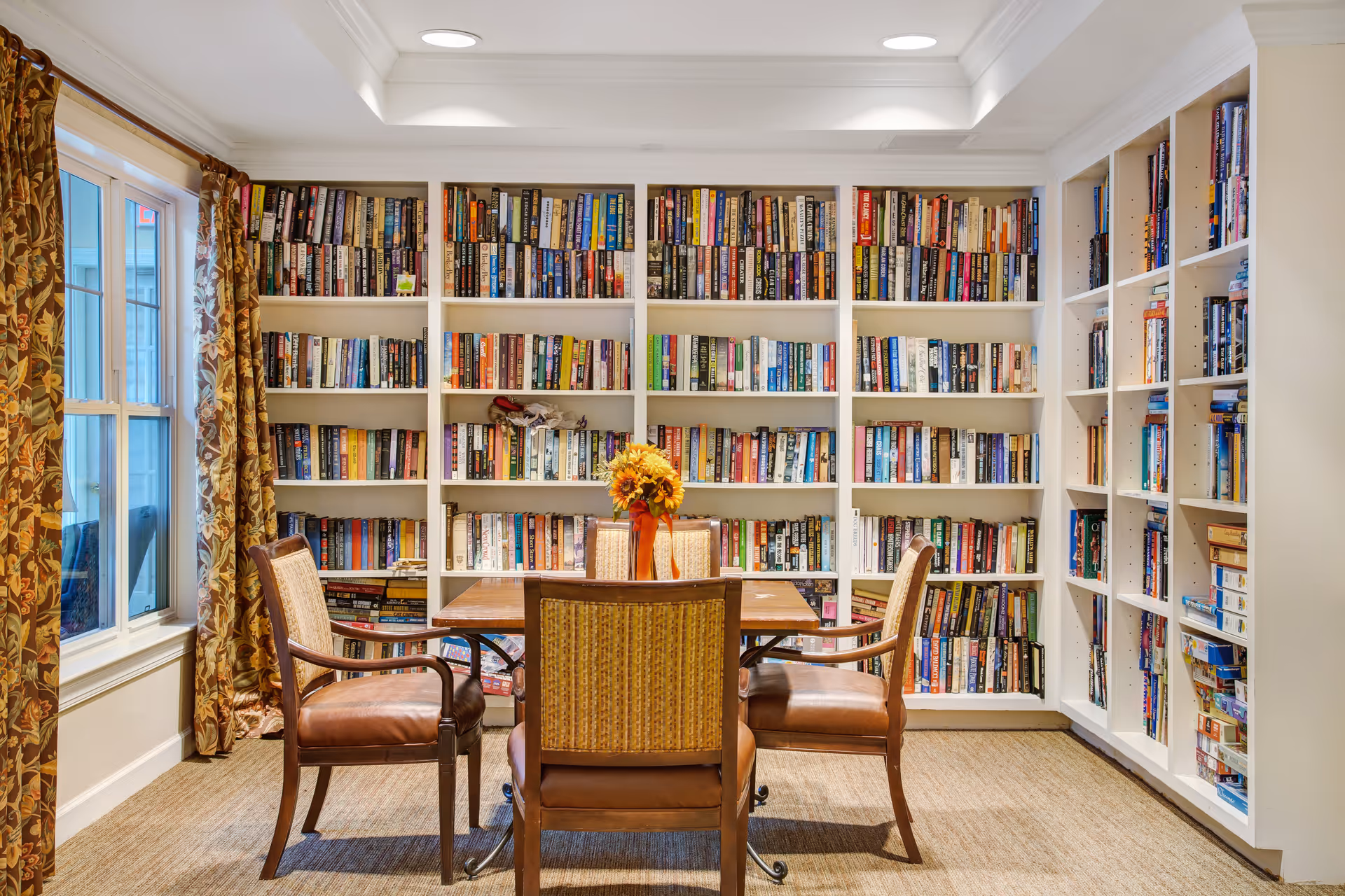 A cozy reading room with a wooden table surrounded by four cushioned chairs. Behind the table, there are white built-in bookshelves filled with a variety of books. A vase with sunflowers is placed on the table. To the left, there is a window with floral patterned curtains allowing natural light into the room.