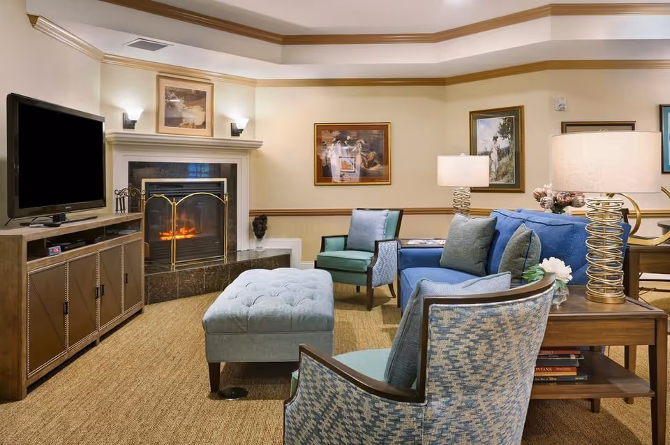 A cozy living room with a lit fireplace, flat-screen TV, blue sofa, patterned armchairs and a tufted ottoman.