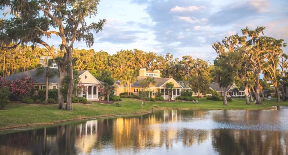Lakeside view of single-story residential buildings and trees reflected in a calm pond at sunset.