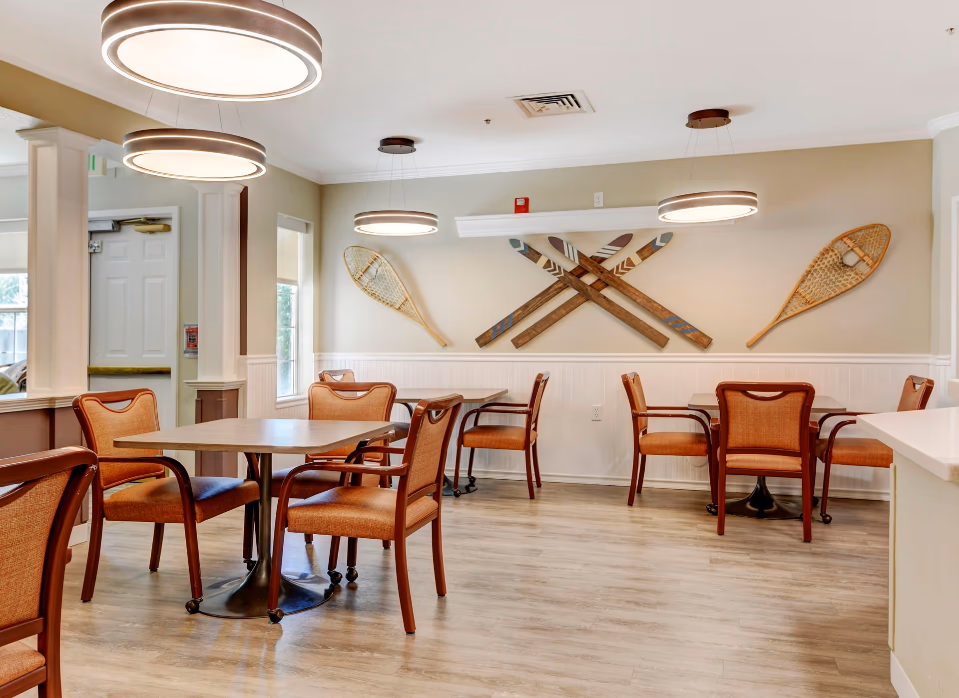 A dining area with several tables and orange cushioned chairs on a light wood floor. The walls are beige with white wainscoting, and there are decorative wooden snowshoes and paddles mounted on the wall. Modern circular pendant lights hang from the ceiling, and there is a window letting in natural light.