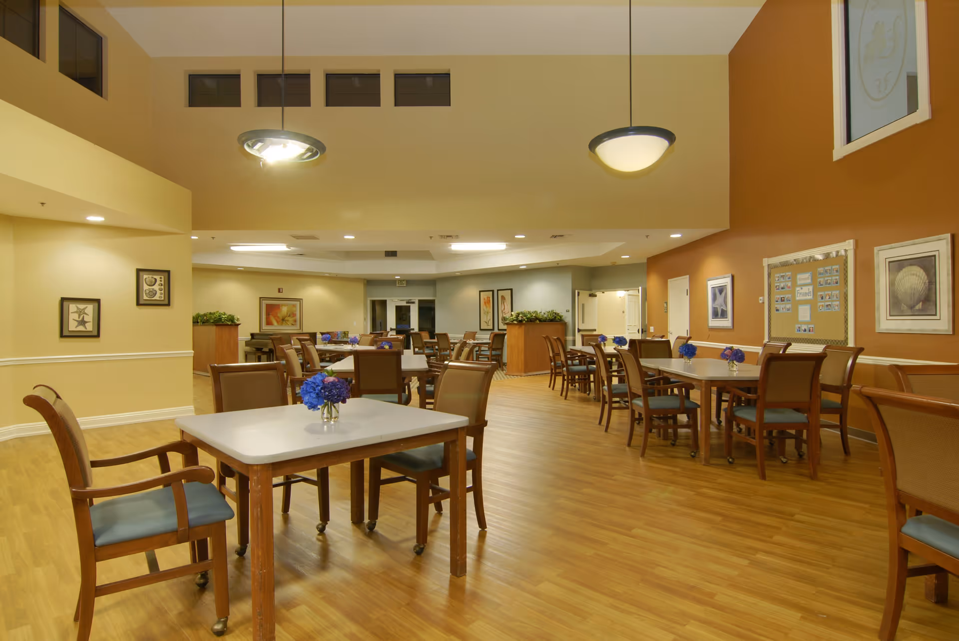 A spacious dining room with multiple wooden tables and chairs arranged neatly. Each table has a small vase with blue flowers. The room has warm yellow and orange walls adorned with framed artwork and bulletin boards. The floor is wooden, and the ceiling has modern light fixtures.