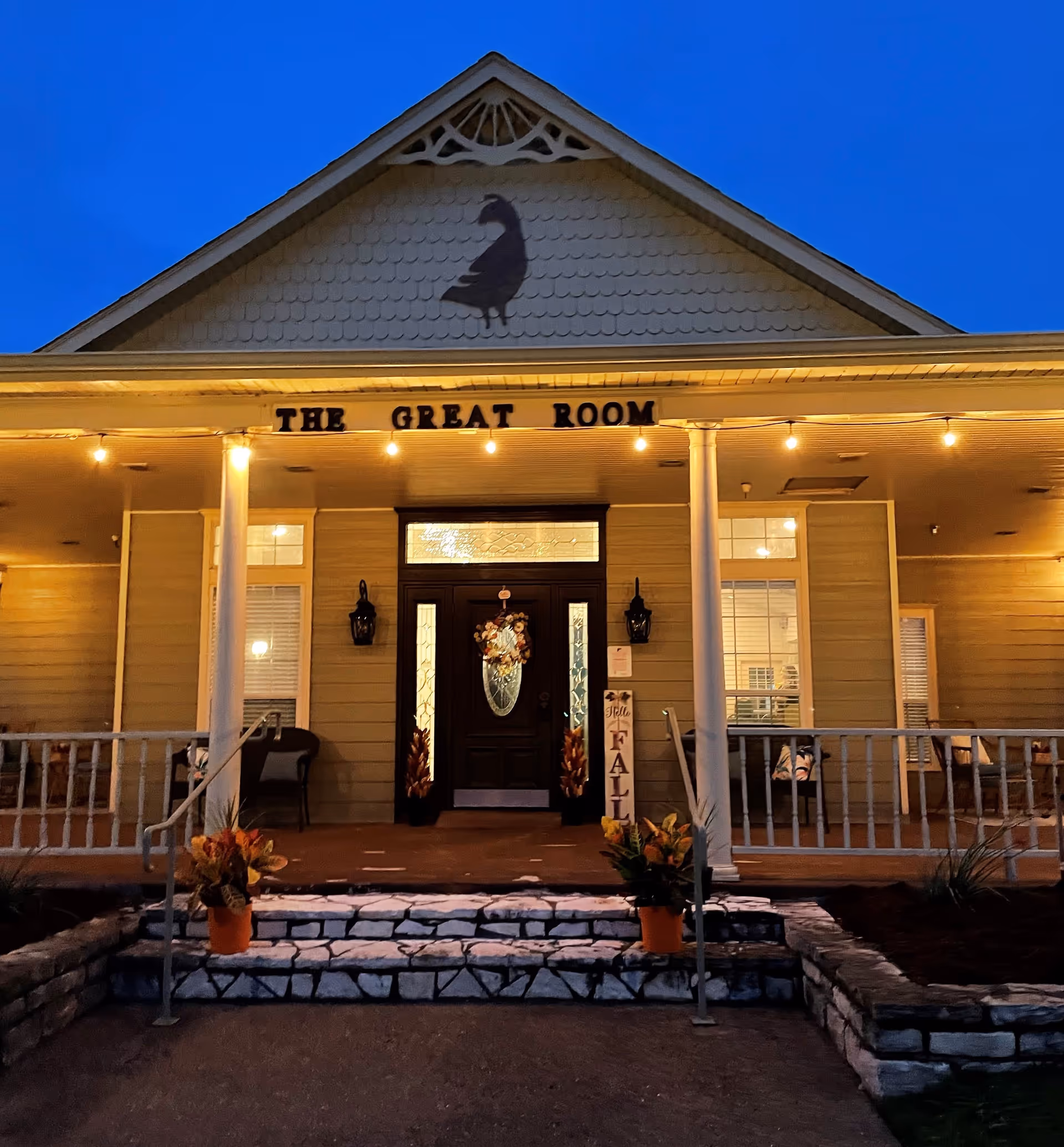 Front entrance of a lit senior living building at dusk with a porch, columns, fall planters, and a sign reading "THE GREAT ROOM" above the door.