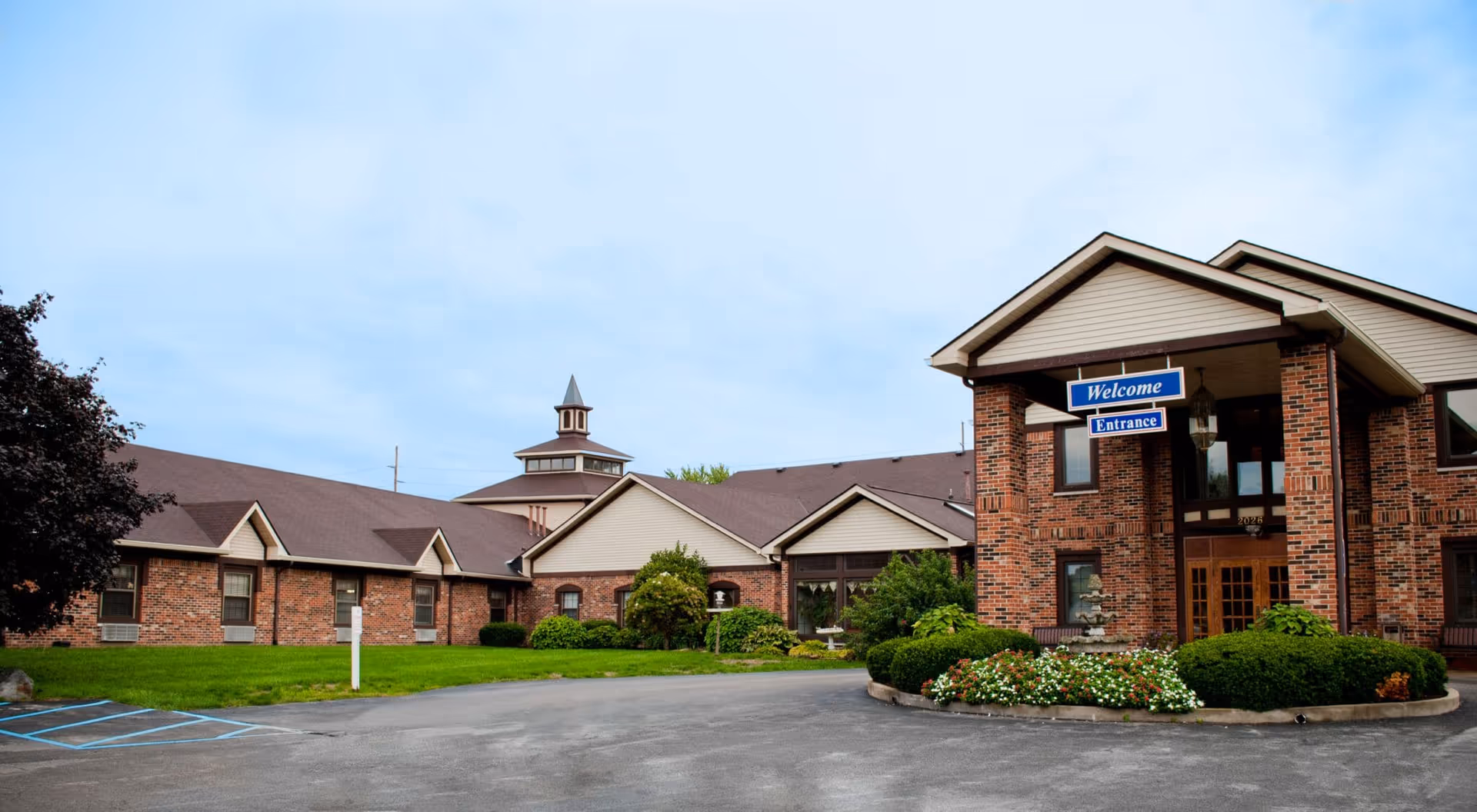 Front exterior of a brick senior living facility with a covered entrance, a 'Welcome Entrance' sign, and landscaped grounds.