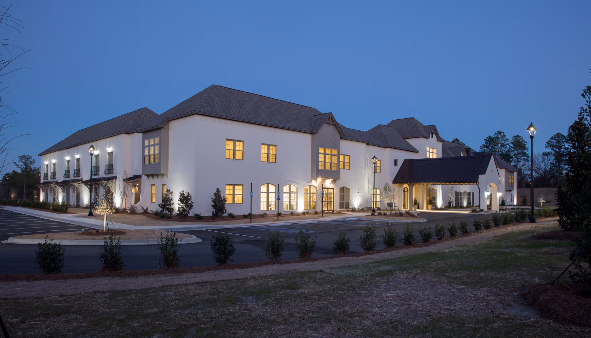 Exterior view of a large, two-story senior living facility building at dusk with lights on inside and outside. The building has a white facade, multiple windows, a covered entrance, and surrounding landscaping including small bushes and trees. The sky is clear and blue.