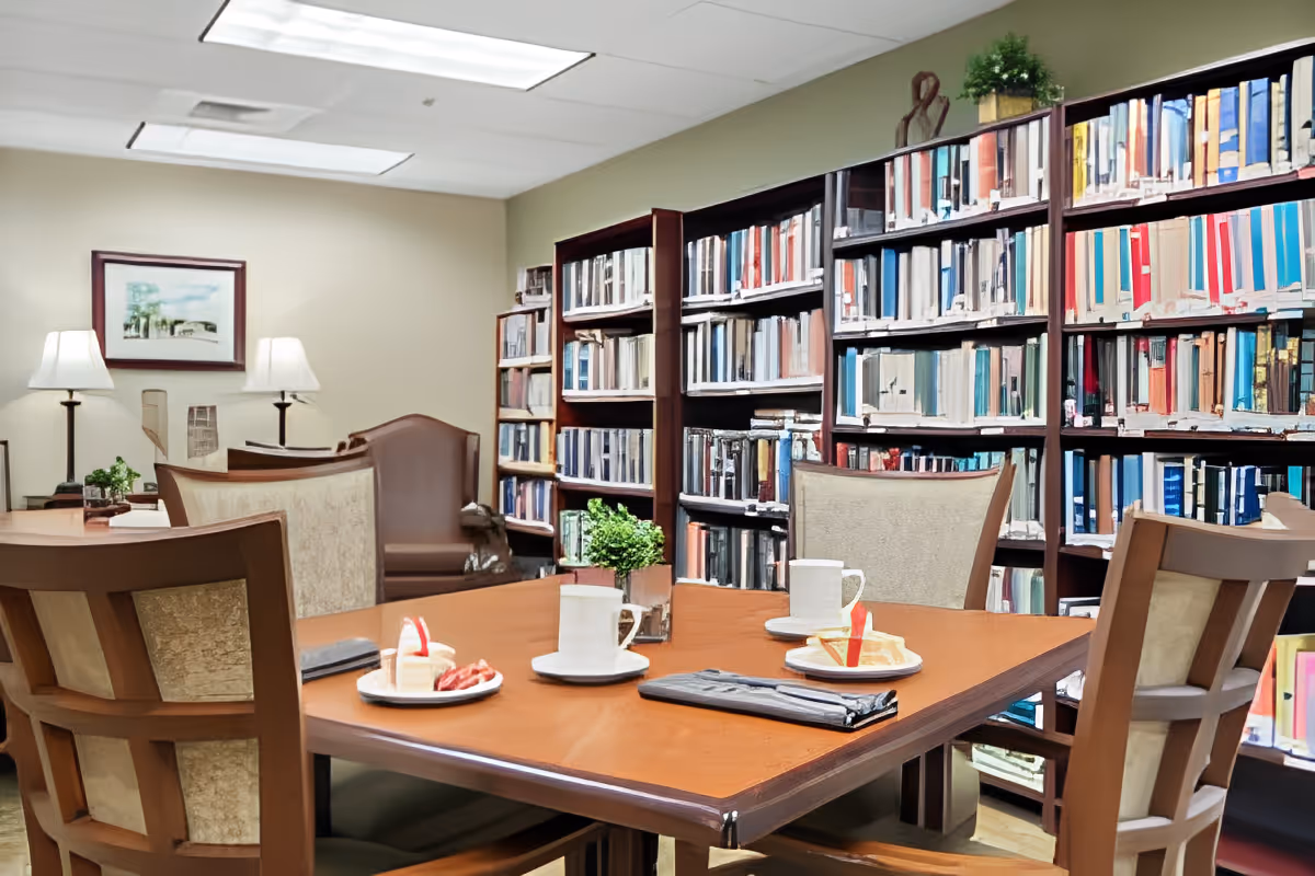 A cozy library room in Carillon Assisted Living of Hendersonville featuring wooden bookshelves filled with books, a wooden table set with plates of dessert and cups, surrounded by cushioned chairs. The room is softly lit by two table lamps and has a framed picture on the wall.
