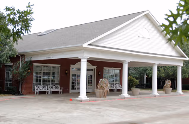 Front exterior view of a single-story brick building with a large white portico supported by columns. There are benches and decorative planters near the entrance, with trees and greenery around the building.