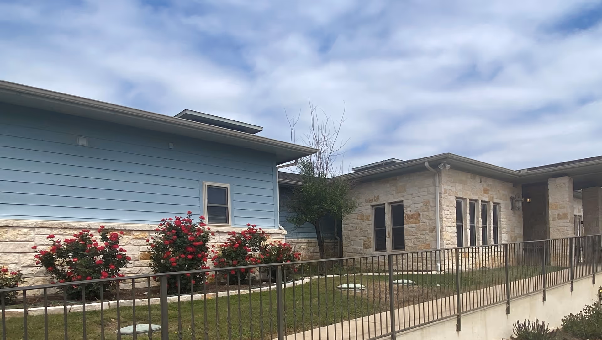 Exterior view of a single-story building with light blue siding and stone walls, a small garden with red flowers, a tree, and a metal railing along a walkway under a partly cloudy sky.