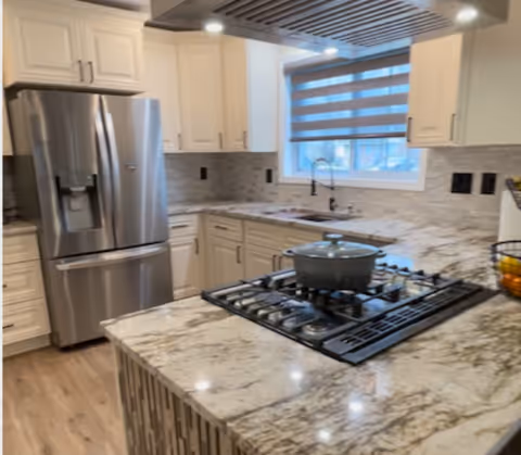 Modern kitchen with white cabinets, a stainless steel refrigerator, a gas stove with a pot on it, marble countertops, a window with a striped blind, and a sink with a black faucet.