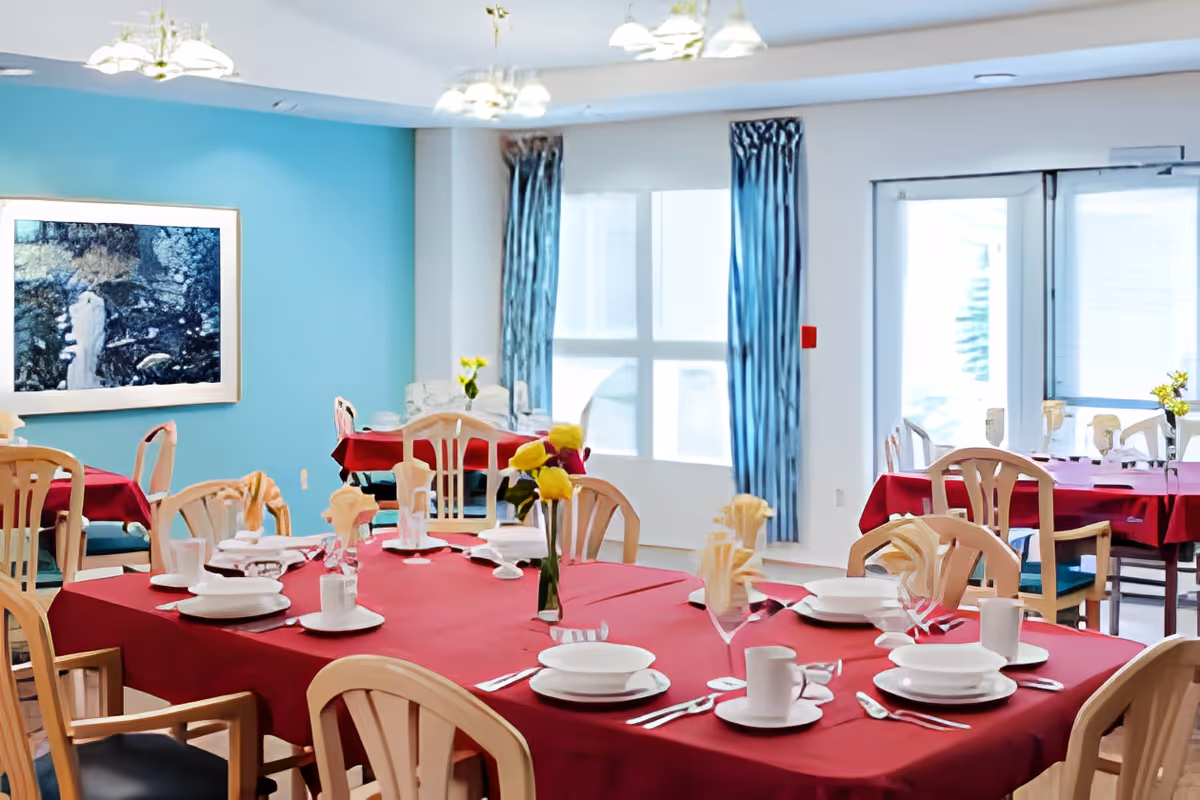 Dining room with several tables covered in red tablecloths, set with white plates, cups, silverware, and napkins. The room has light blue walls, large windows with blue curtains, and a framed artwork on the wall.