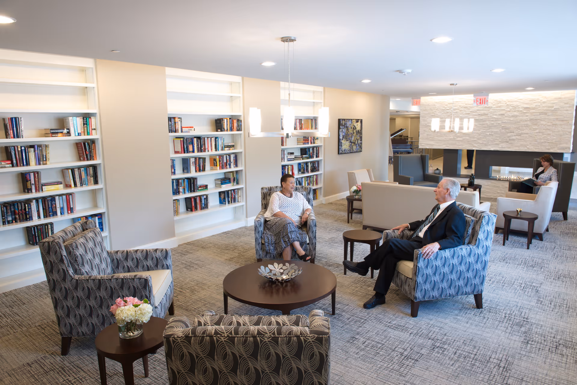 A bright and spacious lounge area with patterned armchairs and round wooden tables. Two elderly people are seated and conversing, while another person reads in the background near a modern fireplace. Bookshelves filled with books line the wall, and contemporary light fixtures hang from the ceiling.