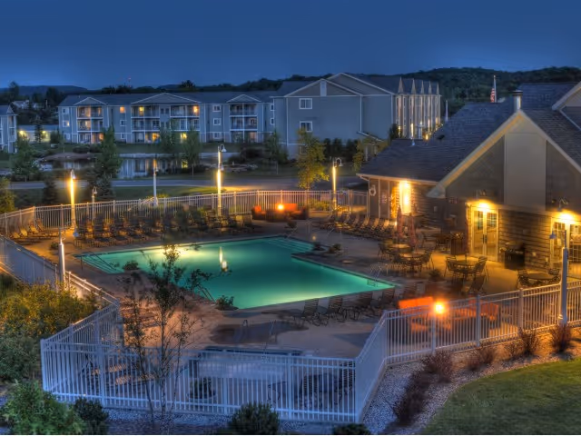 Nighttime view of a well-lit outdoor swimming pool area surrounded by lounge chairs and tables with umbrellas. The pool is enclosed by a white fence, and there is a building with warm lights on the right side. In the background, there are multi-story residential buildings and trees under a clear evening sky.