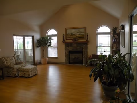 Spacious common room with vaulted ceiling, a central fireplace between arched windows, seating, sliding glass doors, and potted plants on hardwood floors.