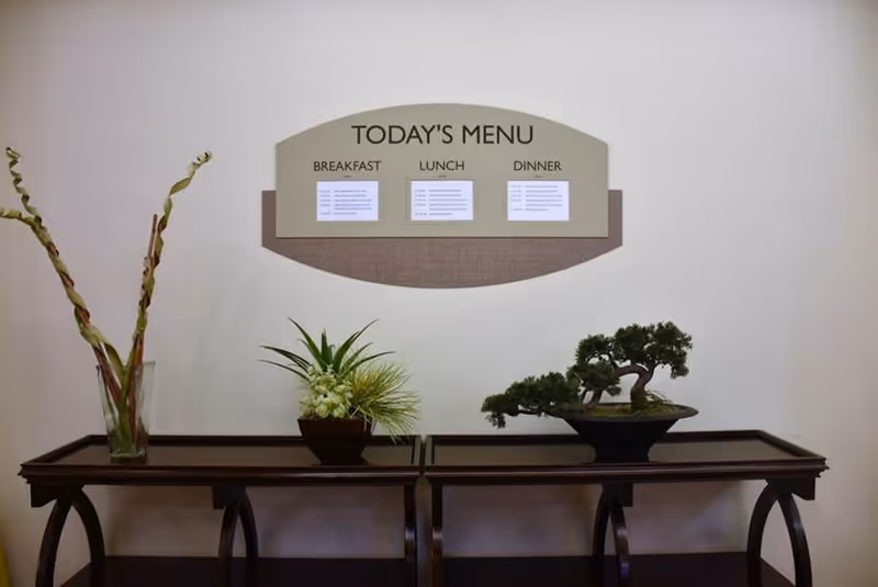 A wall-mounted menu board titled 'Today's Menu' displaying breakfast, lunch, and dinner options above two dark wooden tables. On the tables are decorative plants, including a bonsai tree and a vase with tall twisted branches.