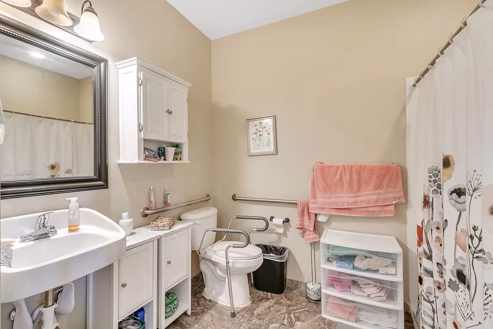 A bathroom with a white sink and mirror on the left, a white cabinet above the toilet, and a toilet equipped with safety rails. There is a pink towel hanging on a rail above a plastic drawer unit on the right side. The shower curtain has a floral pattern, and the floor has a marble-like design.