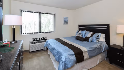 A neatly made bed with blue and brown bedding in a bedroom. The room has a window with vertical blinds, a dresser with a lamp and plant, a nightstand with a lamp, and a decorative 'WELCOME' sign on the air conditioning unit below the window.