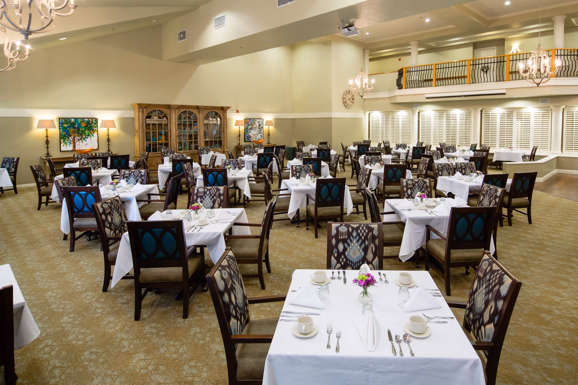 A spacious dining room with multiple tables covered in white tablecloths, each set with cups, plates, silverware, and small floral centerpieces. The room features patterned chairs, chandeliers hanging from the ceiling, a wooden cabinet against the wall, and large windows with white shutters.