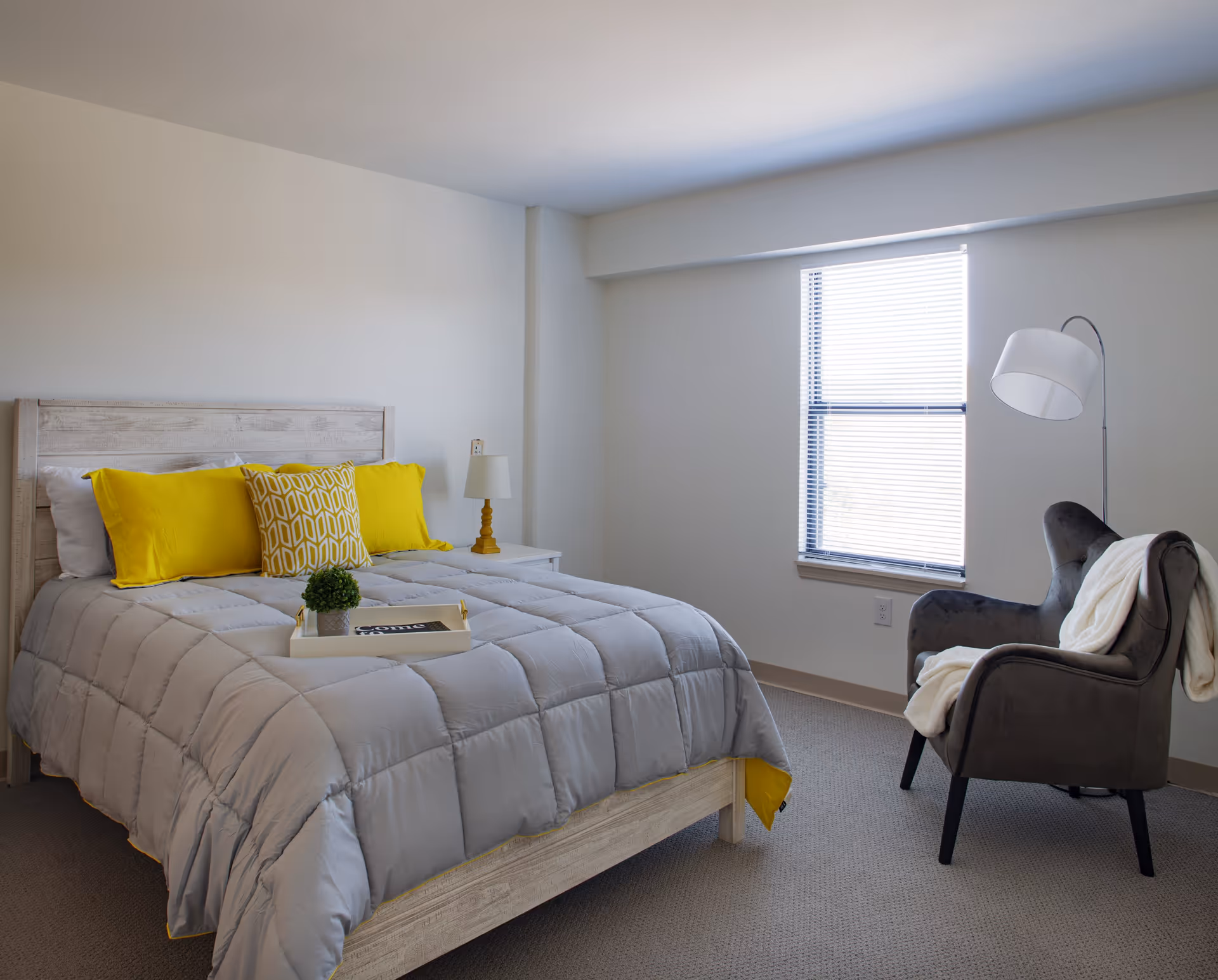 A bedroom with a light wooden bed frame, gray comforter, and bright yellow pillows. There is a white nightstand with a small lamp next to the bed. A gray armchair with a white throw blanket is positioned near a window with blinds. The room has neutral-colored walls and carpeted floor.