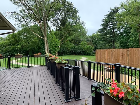 A wooden deck with black metal railings and flower pots containing pink and red flowers. The deck overlooks a green outdoor area with trees, grass, a wooden fence, and a winding pathway under an overcast sky.